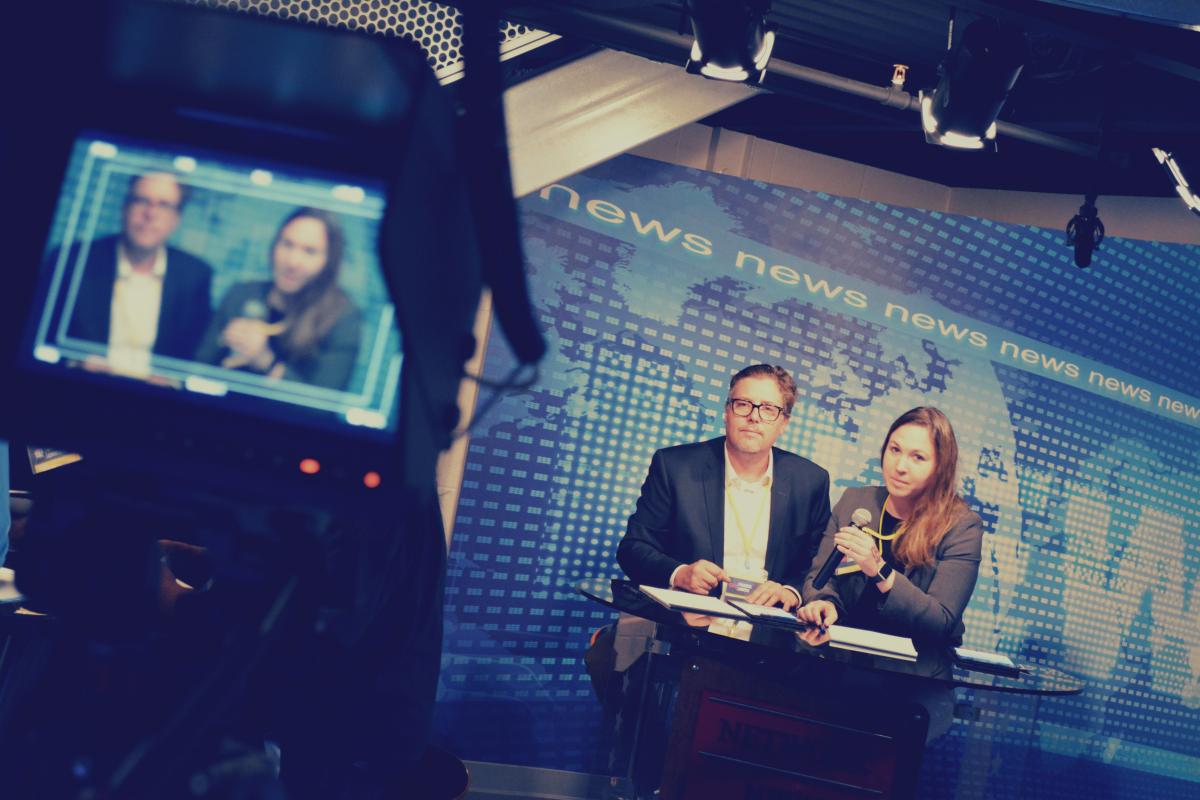 Participants in press roles performing a live news broadcast to all television screens in the Situation Room Experience: Leadership Challenge.